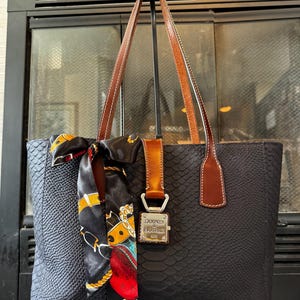 May include: Black tote bag with a snakeskin texture, brown leather handles, and a decorative scarf. The bag features a metal clasp with the brand name "Dooney & Bourke". The scarf has a colorful pattern with gold and red accents.