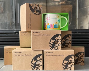 May include: A stack of brown cardboard boxes with the Starbucks logo and the text "You Are Here Collection". The top box has a white ceramic mug with the text "Florida" and a colourful design.