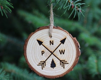 May include: A wooden Christmas ornament shaped like a tree stump with a compass design. The ornament is decorated with a compass rose and the letters N, W, E, and S. The ornament is hanging from a tree branch.
