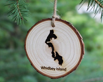 Könnte beinhalten: Ein hölzerner Weihnachtsschmuck mit der Form der Insel Whidbey. Die Inselform ist dunkel gegen das helle Holz. Der Text "Whidbey Island" ist darunter eingraviert. Eine Schnurschlaufe ist zum Aufhängen befestigt.