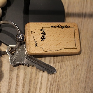 May include: A wooden keychain with the outline of Washington state and the word "washington" engraved on it. A silver key and a beaded wooden strand are also in the image.