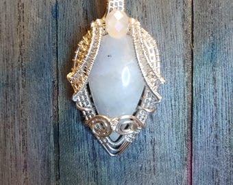 May include: A handcrafted pendant featuring a large, oval, pale blue gemstone, encased in intricate silver wirework. A smaller, faceted, clear gemstone sits above the larger stone. The pendant hangs from a black cord.