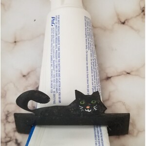 Peut inclure: Distributeur de dentifrice en m&eacute;tal noir en forme de chat avec des yeux verts. Le distributeur contient un tube de dentifrice blanc avec du texte noir.