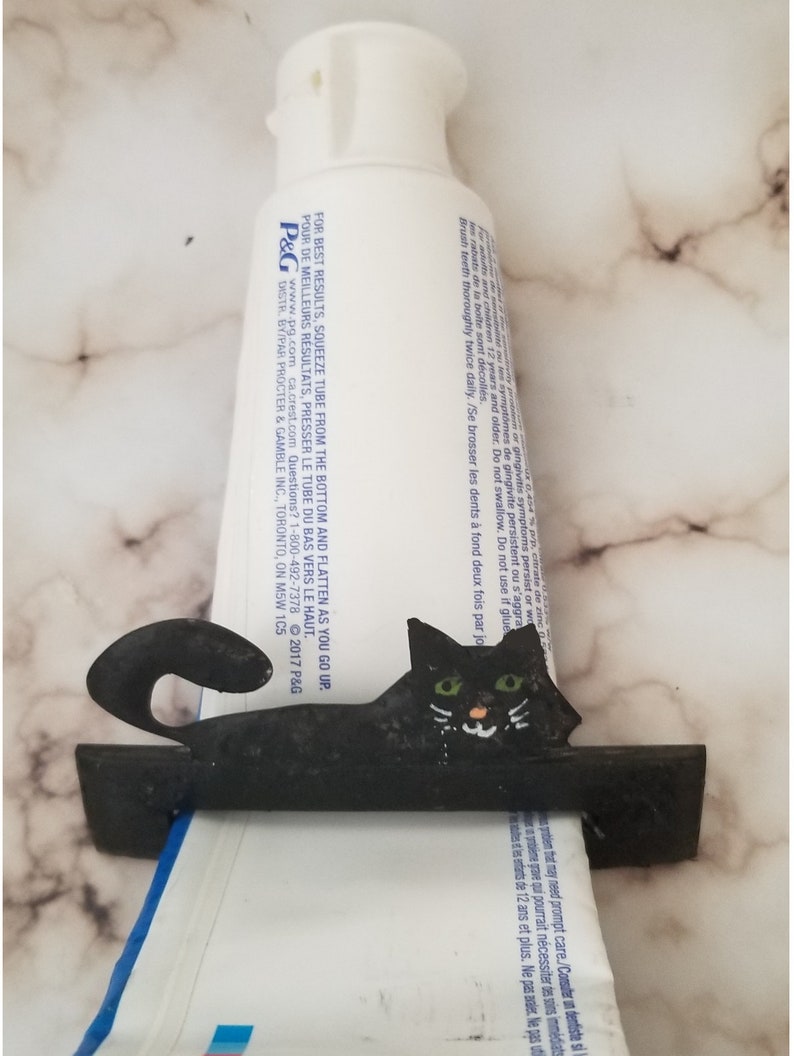 Peut inclure: Distributeur de dentifrice en m&eacute;tal noir en forme de chat avec des yeux verts. Le distributeur contient un tube de dentifrice blanc avec du texte noir.