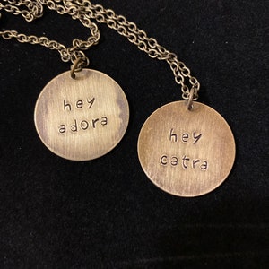 May include: Two gold necklaces with round pendants. The pendants have the words "hey adora" and "hey catra" engraved on them.