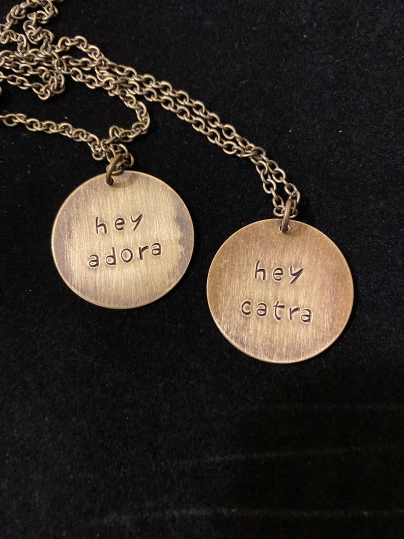 May include: Two gold necklaces with round pendants. The pendants have the words "hey adora" and "hey catra" engraved on them.