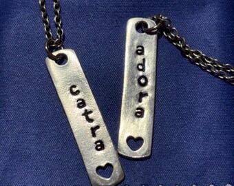 May include: Two silver-toned metal rectangular pendant necklaces on thin chains. Each pendant features vertical stamped lettering, with one reading catra and the other reading adora. Both pendants have a small heart-shaped cutout at the bottom end.