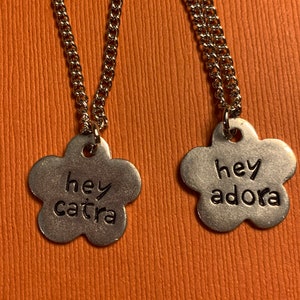 May include: Two silver necklaces with flower-shaped pendants. The pendants have the words "hey catra" and "hey adora" engraved on them.