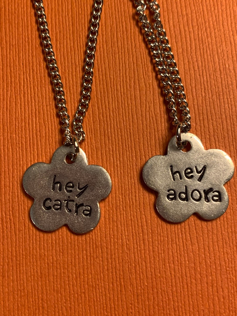May include: Two silver necklaces with flower-shaped pendants. The pendants have the words "hey catra" and "hey adora" engraved on them.