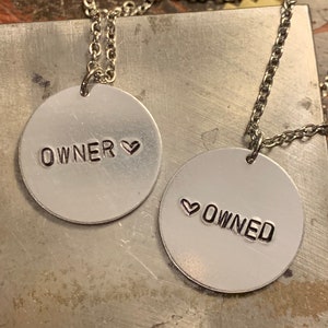 May include: Two silver-colored pendant necklaces with chain. One pendant reads "OWNER" with a heart symbol, the other reads "OWNED" with a heart symbol. The pendants are circular and stamped with black letters.
