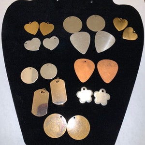 May include: Assortment of metal blanks for jewelry making in various shapes and sizes, including circles, hearts, guitar picks, and dog tags. The blanks are in silver, gold, and copper tones.