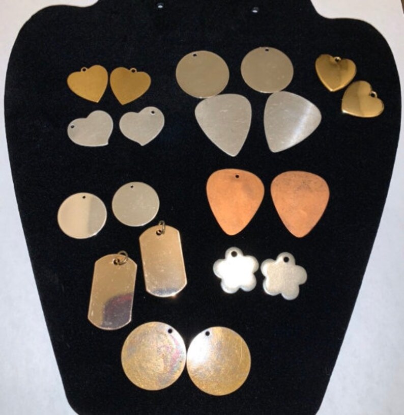 May include: Assortment of metal blanks for jewelry making in various shapes and sizes, including circles, hearts, guitar picks, and dog tags. The blanks are in silver, gold, and copper tones.