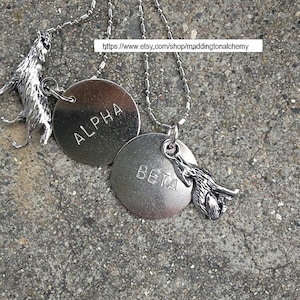 May include: Silver-toned necklaces featuring wolf pendants and engraved circular tags. One tag reads "ALPHA," the other "BETA." The necklaces are displayed on a textured gray surface. The image includes a shop URL.