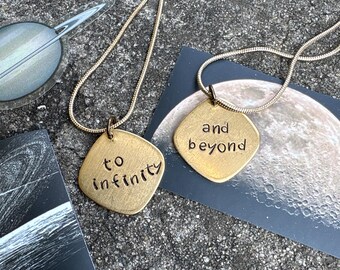 May include: Two gold-toned metal pendants on thin snake chains. One pendant is engraved with the text 'to infinity' and the other with 'and beyond'. The pendants have a soft, brushed finish and a rounded square shape with slightly irregular edges.