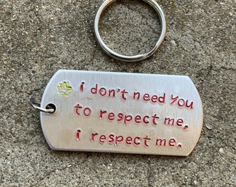May include: A silver metal dog tag style keychain with the text "I don't need you to respect me, I respect me" in red lettering. A yellow starburst design is at the top left. A silver key ring is above the tag.