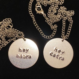 May include: Two silver necklaces with round pendants. The pendants have the words "hey adora" and "hey catra" engraved on them.