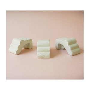 May include: Three off-white, textured, geometric sculptures are arranged on a light pink background. Two sculptures have an arched design with steps, and one is a rectangular block with horizontal ridges. The objects appear to be made of plaster or a similar material.