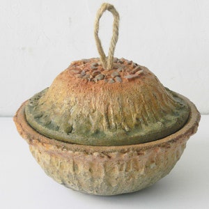 May include: A ceramic lidded bowl with a rope handle. The bowl is decorated with a textured surface and small stones. The bowl is a light brown colour with green and orange accents.