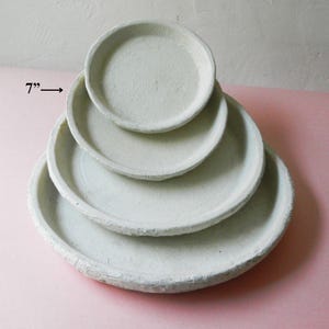 May include: Set of five white ceramic plant saucers, stacked in a pyramid shape. The largest saucer is 17.8 cm in diameter.