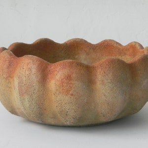 May include: A round, terracotta-coloured planter with a scalloped rim. The planter has a pumpkin-like shape with vertical ridges and a weathered appearance. The colour transitions from a light tan to a reddish-orange at the rim.