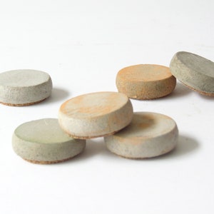 May include: Six round coasters in shades of grey, beige, and green. Each has a textured, concrete-like appearance and a cork base. The coasters are arranged on a white surface. Each coaster measures approximately 8 cm in diameter.