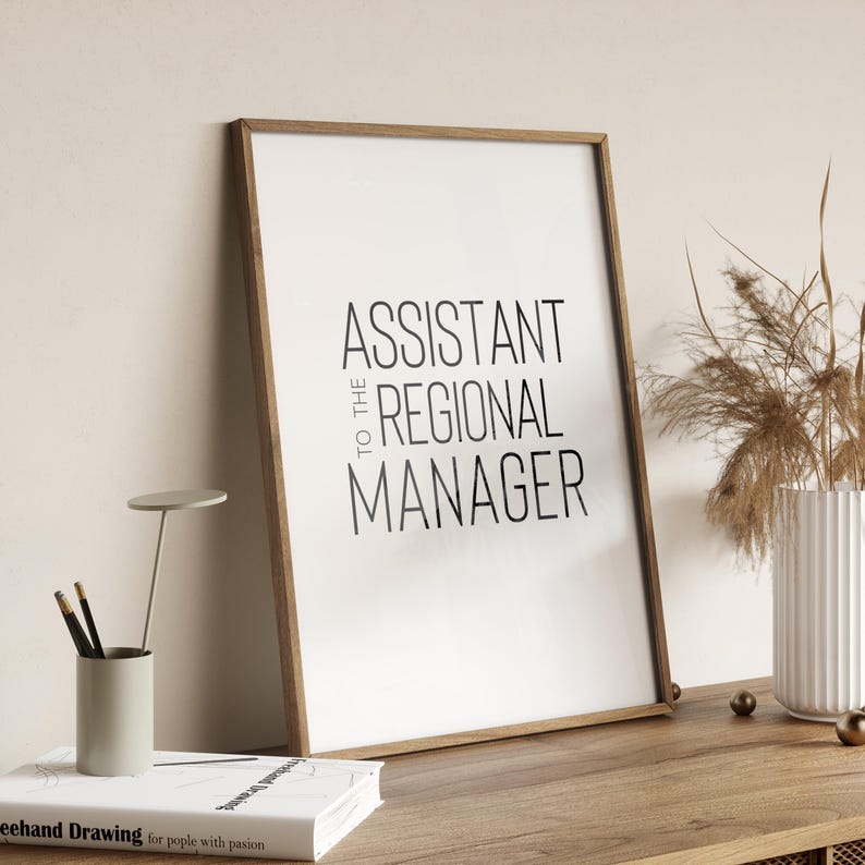 May include: A framed print with the text "Assistant to the Regional Manager" in black text on a white background. The frame is a light brown wood.