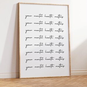 May include: A framed print with the phrase "your mental health matters" repeated in a handwritten style. The print is in a light wood frame and leans against a white wall on a wooden floor.