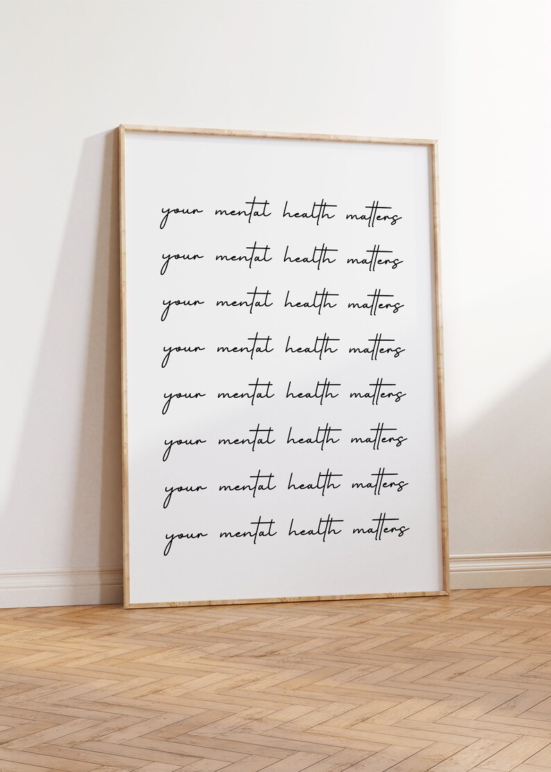 May include: A framed print with the phrase "your mental health matters" repeated in a handwritten style. The print is in a light wood frame and leans against a white wall on a wooden floor.