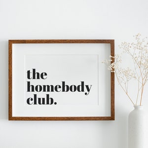 K&ouml;nnte beinhalten: Ein gerahmter Druck mit dem Text "the homebody club." in schwarzer Fettschrift auf wei&szlig;em Hintergrund. Der Rahmen ist aus braunem Holz und der Druck h&auml;ngt an einer wei&szlig;en Wand.