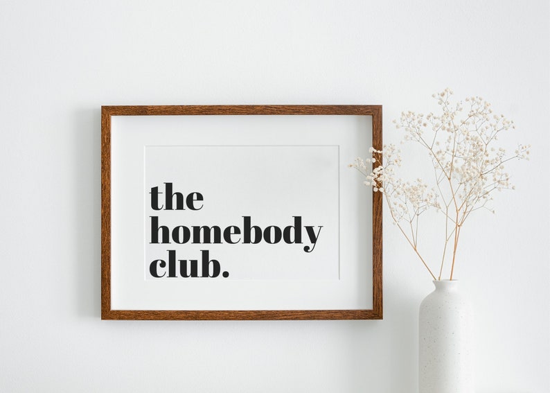 K&ouml;nnte beinhalten: Ein gerahmter Druck mit dem Text "the homebody club." in schwarzer Fettschrift auf wei&szlig;em Hintergrund. Der Rahmen ist aus braunem Holz und der Druck h&auml;ngt an einer wei&szlig;en Wand.