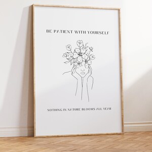 May include: A framed art print with a minimalist line drawing of a person with flowers atop their head. The text reads "BE PATIENT WITH YOURSELF" and "NOTHING IN NATURE BLOOMS ALL YEAR". The frame is light brown.