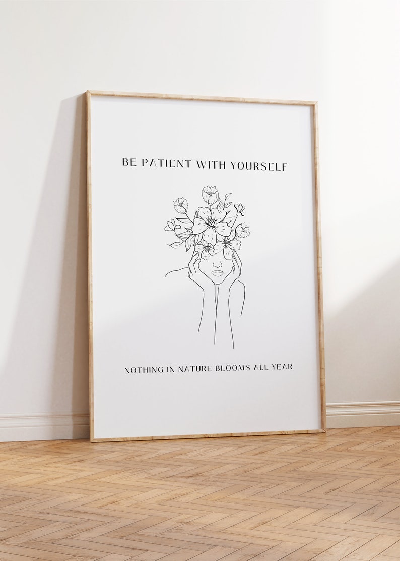 May include: A framed art print with a minimalist line drawing of a person with flowers atop their head. The text reads "BE PATIENT WITH YOURSELF" and "NOTHING IN NATURE BLOOMS ALL YEAR". The frame is light brown.