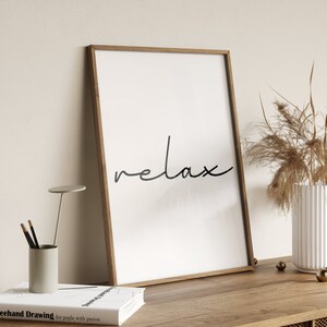 May include: A framed print with the word "relax" written in cursive script on a white background. The frame is a light brown wood.
