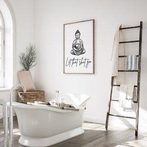 May include: A black and white illustration of Buddha sitting in meditation with the text "Let that shit go" printed below. The illustration is framed and hanging on a white wall in a bathroom setting.