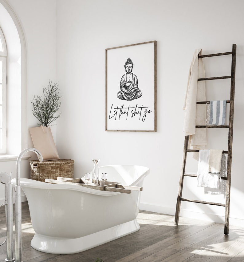 May include: A black and white illustration of Buddha sitting in meditation with the text "Let that shit go" printed below. The illustration is framed and hanging on a white wall in a bathroom setting.