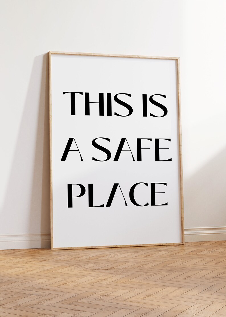 May include: A framed art print with the words "THIS IS A SAFE PLACE" in bold black letters on a white background. The frame is a light wood color, and the print is leaning against a white wall on a wooden floor.