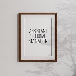 May include: A framed print with the text "Assistant to the Regional Manager" in black lettering on a white background. The frame is brown wood.