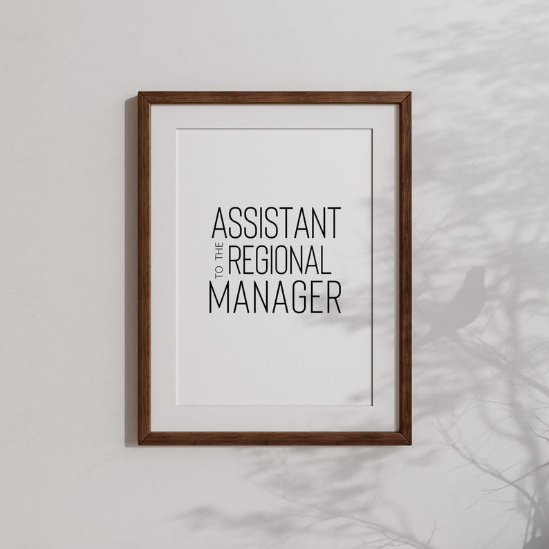 May include: A framed print with the text "Assistant to the Regional Manager" in black lettering on a white background. The frame is brown wood.
