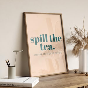 May include: A framed art print with the words "spill the tea" in teal, followed by "YOU'RE IN A SAFE SPACE" in a smaller font. The print has a light peach background and a wooden frame. The artwork is displayed on a wooden surface.