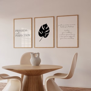 May include: Three framed prints on a white wall above a wooden table with two beige chairs. The prints feature black text and a black silhouette of a leaf. The text on the prints reads "PROGRESS not PERFECTION", "just in case no one told you today hello you matter i value your time i see your effort you are worthy", and a leaf.