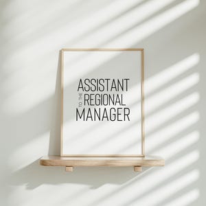 May include: A framed print with the text "Assistant to the Regional Manager" in black lettering on a white background. The frame is light wood and the print is sitting on a light wood shelf.