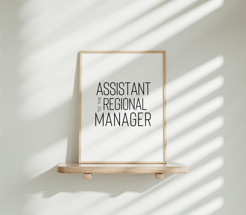 May include: A framed print with the text "Assistant to the Regional Manager" in black lettering on a white background. The frame is light wood and the print is sitting on a light wood shelf.