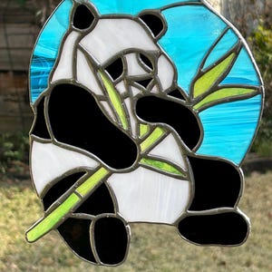 May include: Stained glass panda bear suncatcher with a blue background. The panda is black and white, holding green bamboo. The suncatcher is round and has a silver metal border.