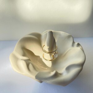 Può includere: Porta anelli in ceramica bianca a forma di fiore con un anello d'oro con una pietra bianca in cima.