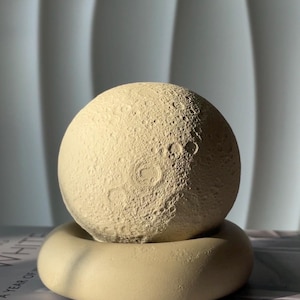 Könnte beinhalten: Eine beige Keramik-Mondskulptur mit einer strukturierten Oberfläche, die der Oberfläche des Mondes ähnelt. Die Skulptur steht auf einem beigefarbenen Keramiksockel.