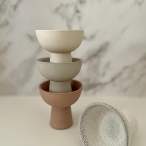 Può includere: Set di quattro ciotole in ceramica in varie tonalità di beige, grigio e bianco. Le ciotole sono impilate l'una sull'altra, con la ciotola più grande in basso e la ciotola più piccola in alto. Le ciotole hanno una superficie testurizzata e una forma leggermente arrotondata.