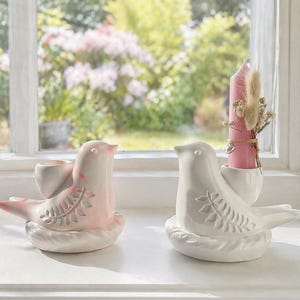 Può includere: Due portacandele a forma di uccello in ceramica. Uno è bianco, l'altro rosa con dettagli bianchi. Ogni uccello è posato su una base a forma di nido e tiene una coppetta per candele. Una candela rosa con fiori secchi è in un supporto.