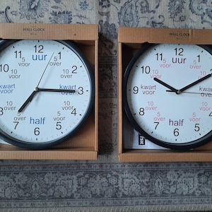 Può includere: Due orologi da parete con cornice nera, quadranti bianchi e lancette nere. Gli orologi hanno i numeri da 1 a 12 e parole olandesi per l'ora, come "kwart voor" e "half".