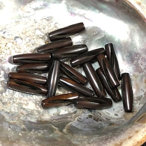 May include: A collection of dark brown, smooth, cylindrical beads, possibly made of wood or bone, arranged in a shell-shaped bowl.
