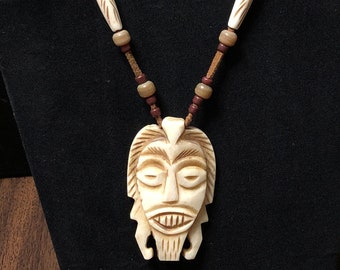 May include: A carved bone pendant necklace with a tribal mask design. The necklace is made with brown and white beads.